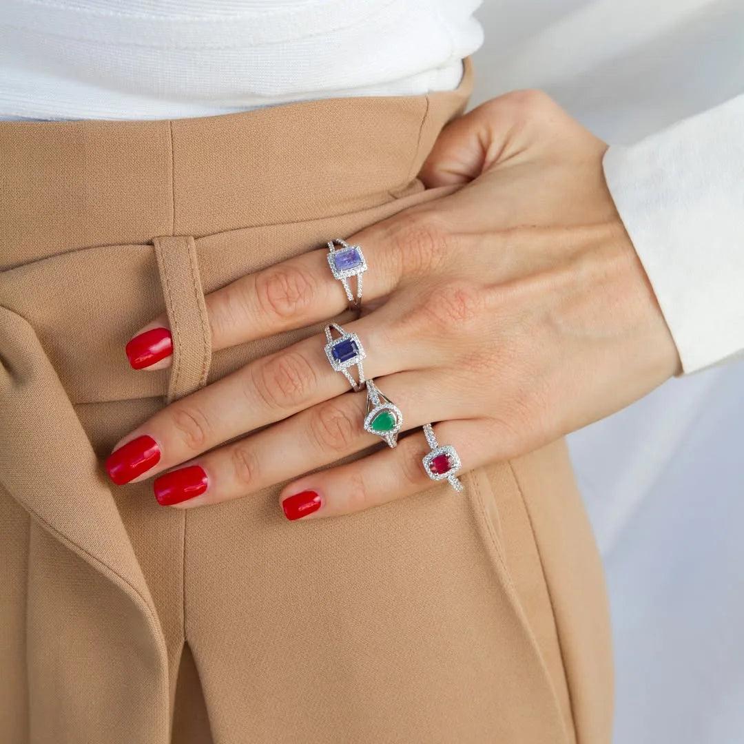 Close-up of hand wearing Cavell gemstone rings with sapphire, emerald, and ruby in sterling silver, styled with tan trousers and red nails.