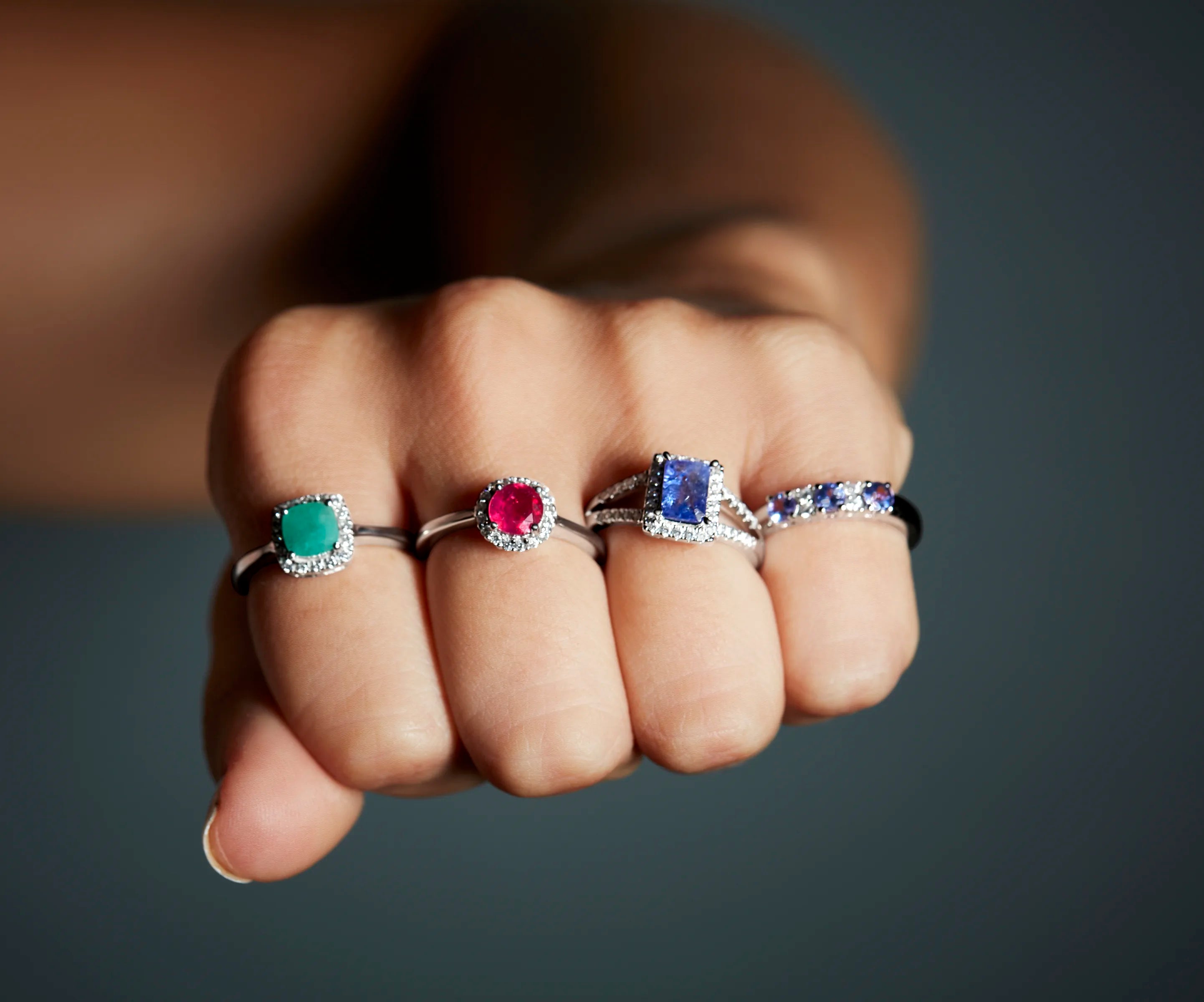 Close-up of a hand wearing Cavell gemstone rings with emerald, ruby, sapphire, and tanzanite on a dark background.